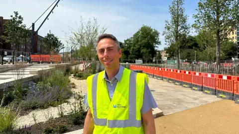 Andrew Spence Councillor Ross is wearing a high vis vest. Hard landscaping is visable in the background with planting and a crane and red barriers.