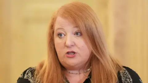 PA Media Naomi Long is wearing a black and white t shirt and a necklace. She has ginger hair. The background is blurry.