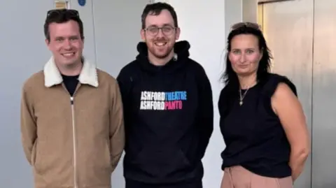 Two men standing next to a woman. One man is wearing a brown jacket and the other is wearing a black hoodie which says "Ashford Theatre. Ashford Panto" on it. The woman is wearing a black shirt with no sleeves.