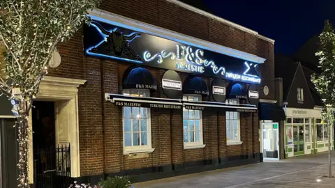 A night time picture of the F&S Turkish restaurant along a wide pedestrianised pavement, there are also trees decorated in fairy lights in raised stone planters. The lettering on the sign is illuminated from behind, other shops can be seen on the left, also with lit signs.