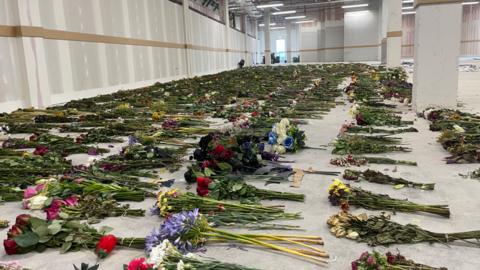 Rows of bouquets of flowers laid out in a large indoor space, suggesting a memorial event