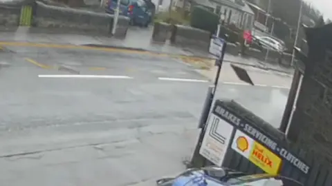 Metal manhole or utility cover lifted into the air beside a roadside sign outside a garage, with wet pavement, a residential street, and light spray or steam visible nearby