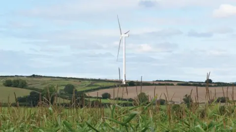 A computer generated image of a tall wind turbine in the English countryside on a sunny day