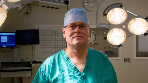 Newcastle Hospitals Professor Francisco Figueiredo, a Consultant Ophthalmologist at Newcastle's Royal Victoria Infirmary. He is wearing his green surgical scrubs and his surgical hat and is in an operating theatre. He is looking at the camera with the theatre lights on above him,