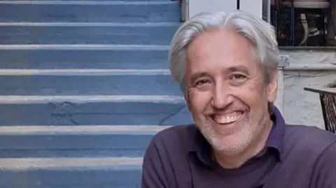 Julian Barwell A man with grey hair and a grey beard and wearing a blue sweatshirt is smiling at the camera.