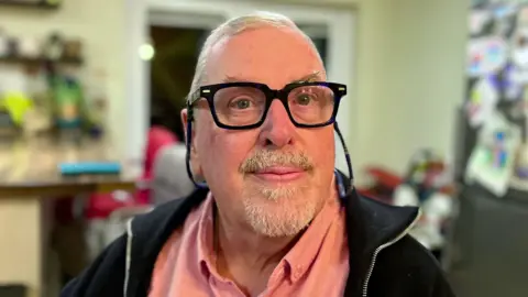 Steve O'Farrell looks directly into a camera. You can see his head and shoulders. He is wearing glasses, a pink shirt and black jacket. The background is blurred, but he appears to be sat in a kitchen.