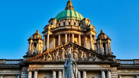 Getty Images A close up of Belfast City Hall with a blue sky. A statue of Queen Victoria is in the centre. The building has numerous pillars and a green dome. 