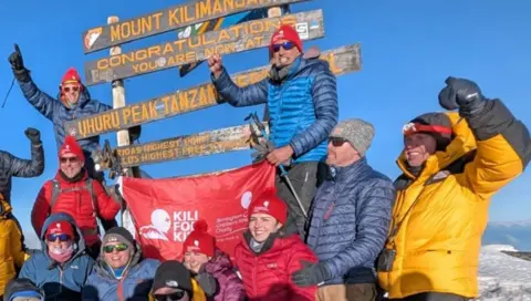 People wearing coats at the top of the mountain, next to a red Kili for Kids banner. Some of the people have an arm outstretched.