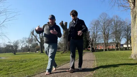 Robert, on the left, wears a cap and jumper with leather elbow patches and light blue trousers. He is not wearing shoes.
Reporter Harry Parkhill, next to him, wearing a bomber jacket, tie and smart trousers is also bare foot. They are walking along a gravel path through a leafy park.