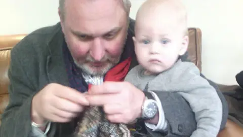 Gary Ray Smith A man with a grey beard and grey hair sits on a brown sofa with knitting needles. He is knitting a baby blanket. He has his arm around a small child sat on his knee who has blue eyes. The baby boy is wearing a grey jumper. 