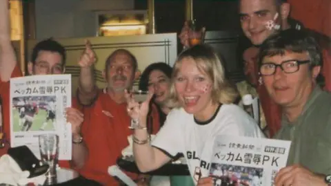 Tom Barnard A group of five people are gathered around a table in a bar or restaurant. Several are holding drinks, and two people are holding up printed pages featuring a football image and Japanese text. One person in the centre foreground is raising a glass towards the camera, while another person in the background has face paint on one cheek and is holding a drink aloft.