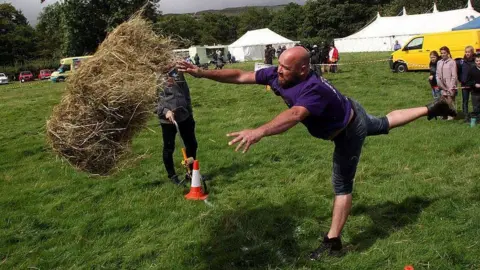 World Alternative Games Man in mid throw, one foot extended behind him, as judge prepares to measure the distance