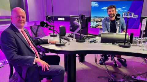 Both Sir Ashley Fox and radio host Charlie Taylor photographed in the BBC Radio Somerset studio, which is lit in purple with a TV screen in the background. They are sat down and facing the camera. 