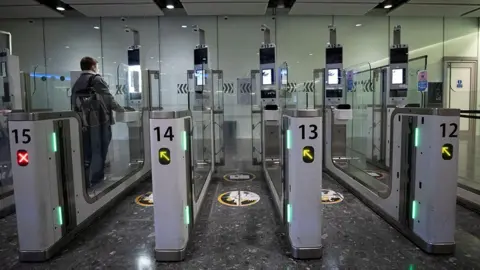 Alamy Passport e-gates in an airport. There are five lanes in the photo and a man stood in one of them. He is holding his passport on the scanner waiting to be let through the gate.
