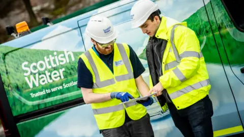 Scottish Water Two Scottish Water workers in high-visibility jackets and hard hats stand next to a Scottish Water van