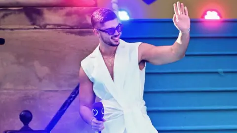 Getty Images Andrew Lambrou wearing a white vest waving to the crowds and holding a microphone at the Eurovision turquoise carpet event in Liverpool