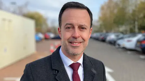 Sam Read/BBC Kevin Bonavia smiling in a car park while smartly dressed. He has short black hair and stubble on his face. He is wearing a white shirt, burgundy tie and a textured black overcoat.