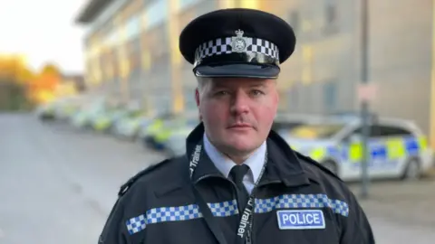 Chief Inspector Ben Huggins stares directly at the camera in full uniform, including a black police hat, black jacket, white shirt and black tie. Behind him is a row of police cars, out of focus. 