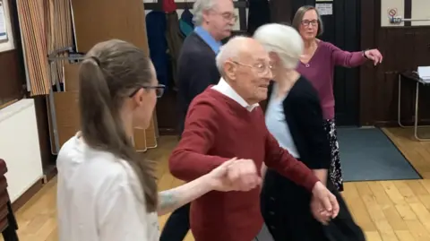 Eddie Gibbs holds the hand of his dance partner who is wearing a white top. The dancing session is taking place in a hall with a wooden floor.