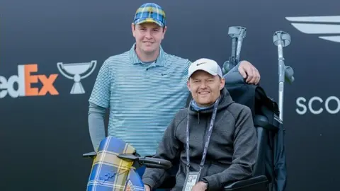 Callum Huntington stands next to Robert McIntyre Scott who is in a wheelchair. Robert is wearing a blue and yellow checkered cap and a striped blue top. Scott is wearing a white Nike cap and gray hoodie.