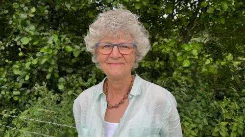 Caroline is wearing a pale green shirt, necklace and glasses. She has grey curly hair and she's standing by a hedgerow