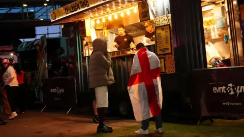Owen Humphreys/PA Two people queuing at a food stand with a sign which reads: "Chicken Ting". 