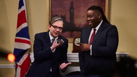 Two actors in a set resembling Ten Downing Street - one is appearing as Keir Starmer, the other as David Lammy, both wearing suits. A Union Flag and painting of Big Ben are behind them