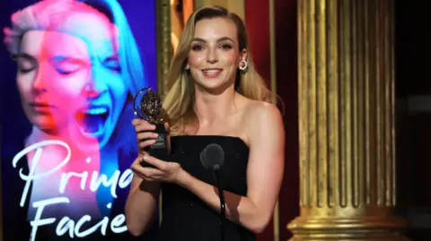 Reuters Jodie Comer pictured smiling and holding a Tony award in front of a poster for Prima Facie. She has long blonde hair and is wearing a black dress and hooped diamond earrings.