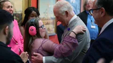 PA Media King Charles hugs 15-year-old Florence McGrellis, who is wearing a pink dress and pink ear defenders.
