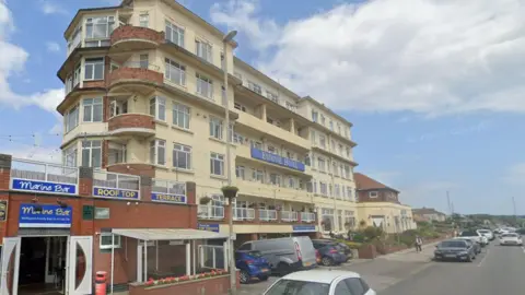 A large six-story hotel with cream walls and art deco features, including curved, red-brick balconies, stands by a road in a seaside town with cars parked outside. To the left is a single-story red-brick bar with roof terrace.