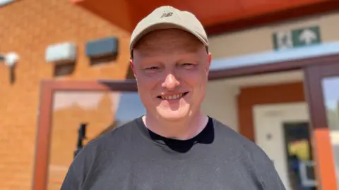 A man is wearing a grey cap and a dark T-shirt. He is smiling and standing in front of a brick wall and an open glass window or door.