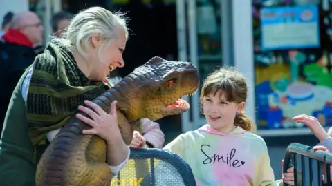 A young girl smiles as a woman holds up a small animatronic dinosaur.  