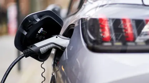 A close up image of a silver and black coloured electric vehicle charger which is plugged into a silver car. The car is out of focus but you can see the rear of the vehicle and the back headlights. 