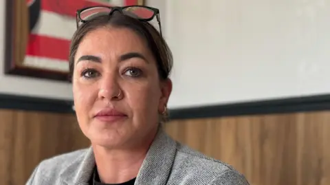 Natasha sitting indoors, wearing a grey blazer over a black shirt. She has dark hair pulled back from her ace. There is a pair of glasses resting on top of her head. She has a slim silver nose ring. She is looking directly at the camera with a neutral expression. The background includes wooden panelling.
