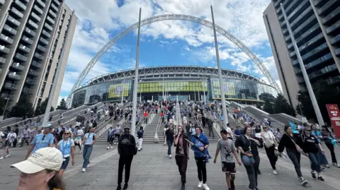 Hundreds of Albion fans leaving Wembley stadium. The photo is taking from the base of the steps, looking up towards the stadium. People walk towards the camera.