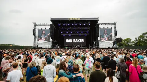 @khaliphotography A large crowd is gathered in front of the main stage at Forwards Festival in Bristol. On the big screens on either side of the stage a main is seen playing a guitar and singing.