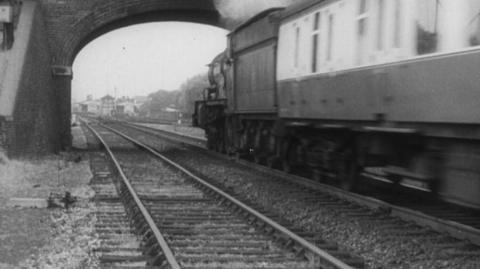 Black and white image of train about to go under a bridge.