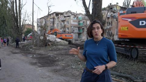 Katy Watson speaking to camera while standing in front of building severely damaged by Russian strike
