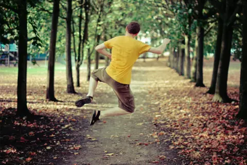 Dave Jones A man in a yellow t-shirt jumps while walking along a path covered with autumn leaves