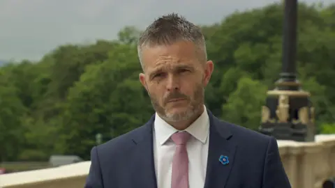 Butler is outside. he's wearing a navy suit with a white shirt and a pink tie. He has a blue floral pin on his jacket.