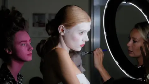 Models stand and wait as they have full face make up applied by a makeup artist