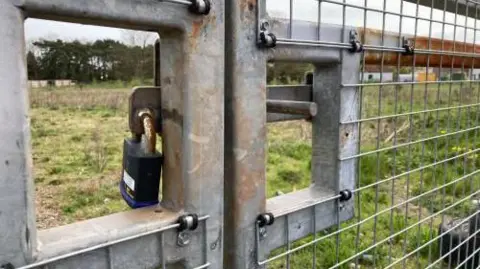 A locked gate with a rusted padlock takes up the foreground. Through the steel railings you can see an empty, unattended lot. 