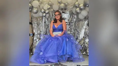 Jessica, who has long dark hair and glasses, smiles as she wears a purple prom dress while sitting in the back of her dad's removals van. There is a silver sparkly backdrop and silver and white balloons behind her.