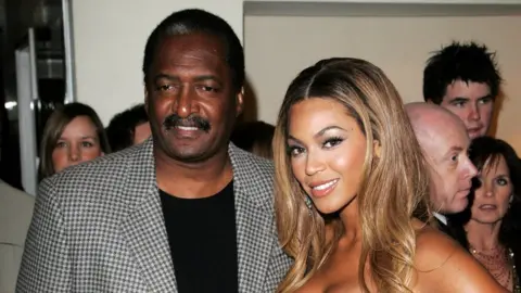 Getty Images Dr Matthew Knowles pictured with Beyoncé at the Dream Girls in 2007. Dr Knowles, a black man in his 70s, has short black hair, brown eyes and a moustache. He wears a black and white dog-tooth blazer over a black top. Beyoncé is a black woman who in this picture would have been in her mid 20s. She has long honey-coloured hair and smiles at the camera. A crowd of people are pictured behind the pair