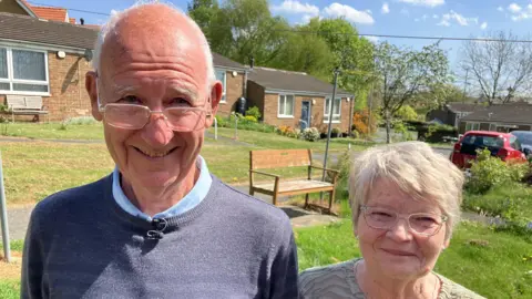 An elderly man pictured standing alongside his wife in a garden. They both wear glasses and are smiling. It is a sunny day and there are houses behind them.