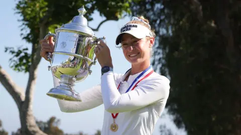 Nelly Korda, wearing a long-sleeved white top and white visor and with a gold medal on a red, white and blue ribbon around her neck, smiles and poses with the silver trophy, holding it up to her right