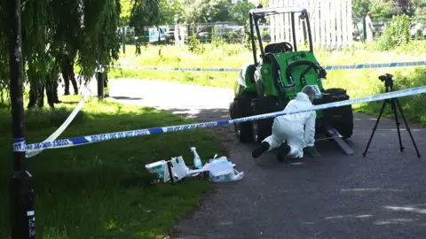 A person in white overalls is on the ground at the front of a small, green, single-seater, four-wheeled vehicle which has two forks at the front. It has a roof over the driver's seat. It is cordoned-off on a path, and has blue and white police tape around trees.