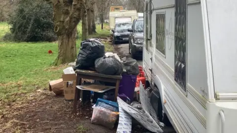 A white caravan parked on a road with a tree lined grass verge to the side. Outside the caravan is a pile of rubbish on and around a wooden table.