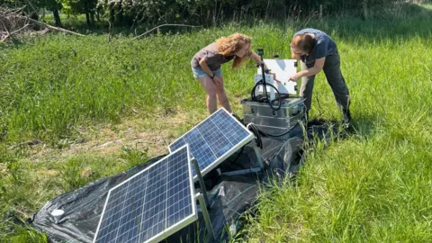 UK Centre for Ecology and Hydrology Two people surround a contraption with solar panels in lush grassland
