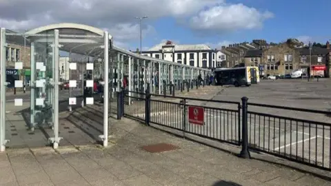 BBC/Katie Radley A long bus shelter with a railing in the foreground and a concrete area behind with a bus in the distance. There are buildings in the background.